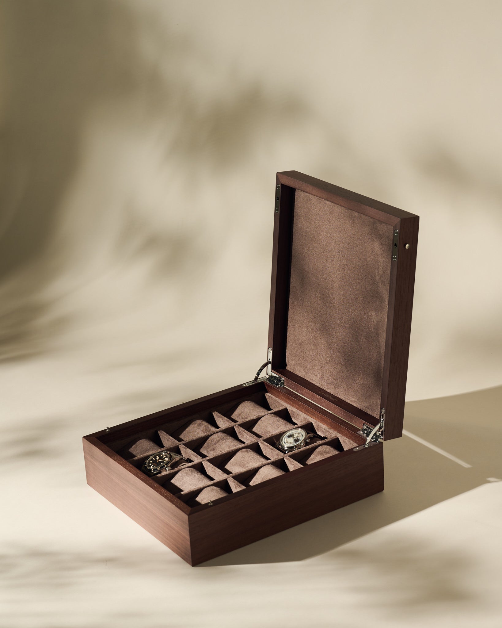 The TAWBURY Grove 12 Slot Wooden Watch Box in dark walnut wood with a solid lid showcases multiple compartments, elegantly organizing two wristwatches and a bracelet on a beige surface with soft shadows.