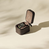 A TAWBURY Fraser 1 Watch Case in brown holds a wristwatch with a black dial, displayed open on a beige surface and casting soft shadows.