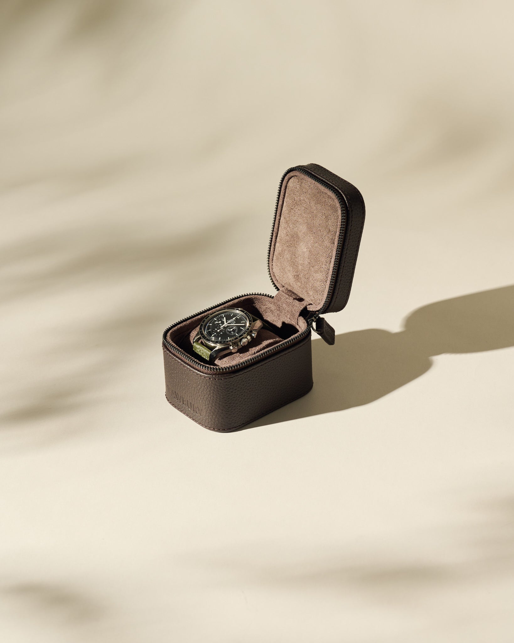 A TAWBURY Fraser 1 Watch Case in brown holds a wristwatch with a black dial, displayed open on a beige surface and casting soft shadows.