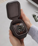 A person holds an open TAWBURY Fraser 1 Watch Travel Case - Brown (Coming Soon), displaying a wristwatch with a brown leather strap and black dial; a green strap and tool rest on the table, showcasing stylish watch protection.