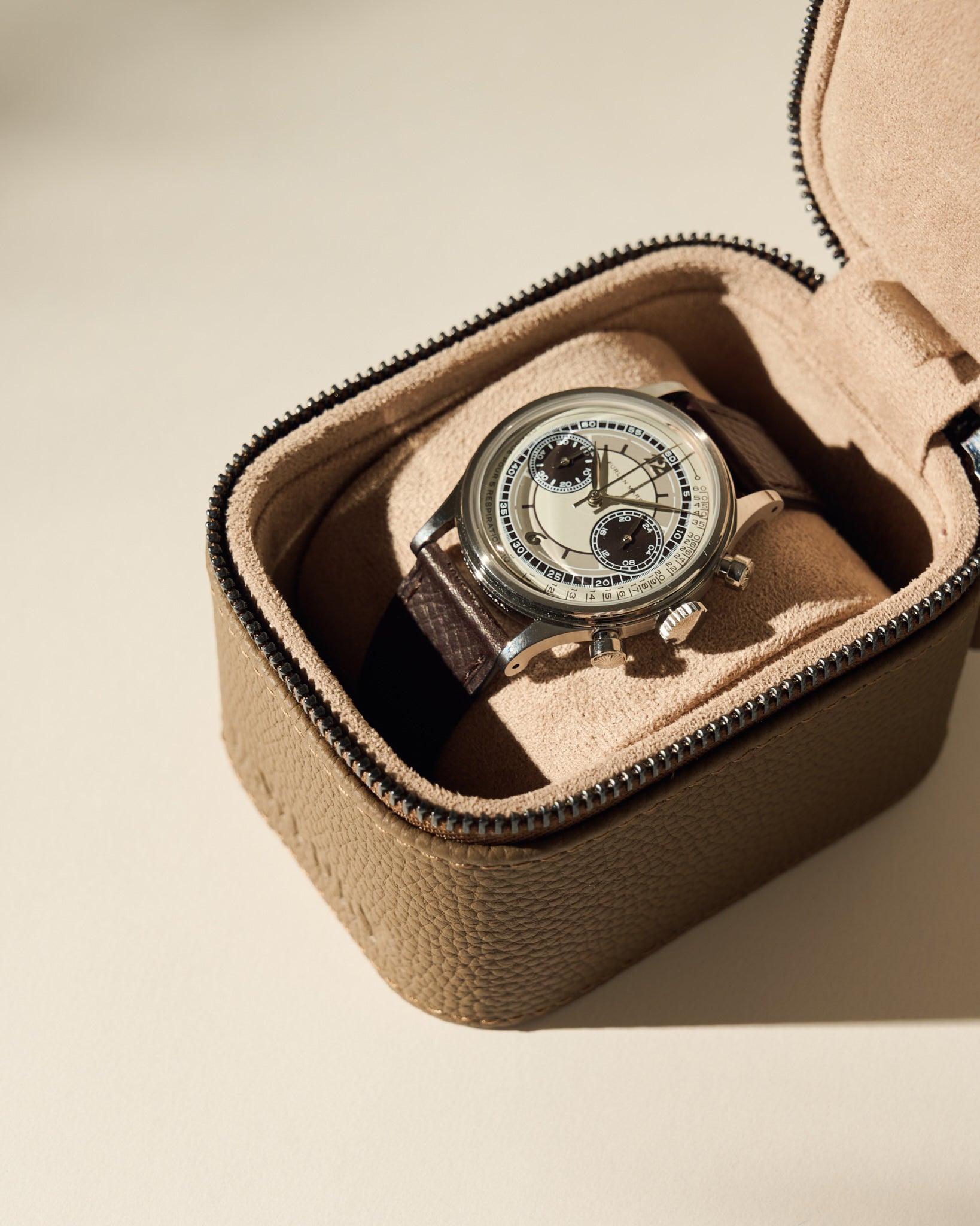 A wristwatch with a brown leather strap and silver casing is displayed inside the TAWBURY Fraser 1 Watch Case - Taupe, featuring a soft suede interior.
