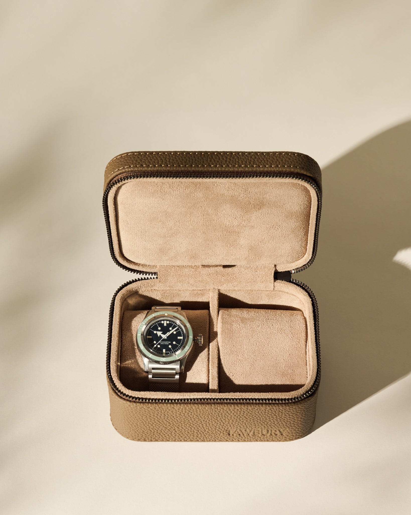 A silver wristwatch with a black face is placed inside the TAWBURY Fraser 2 Watch Case in taupe, a brown leather case featuring suede lining and two compartments.