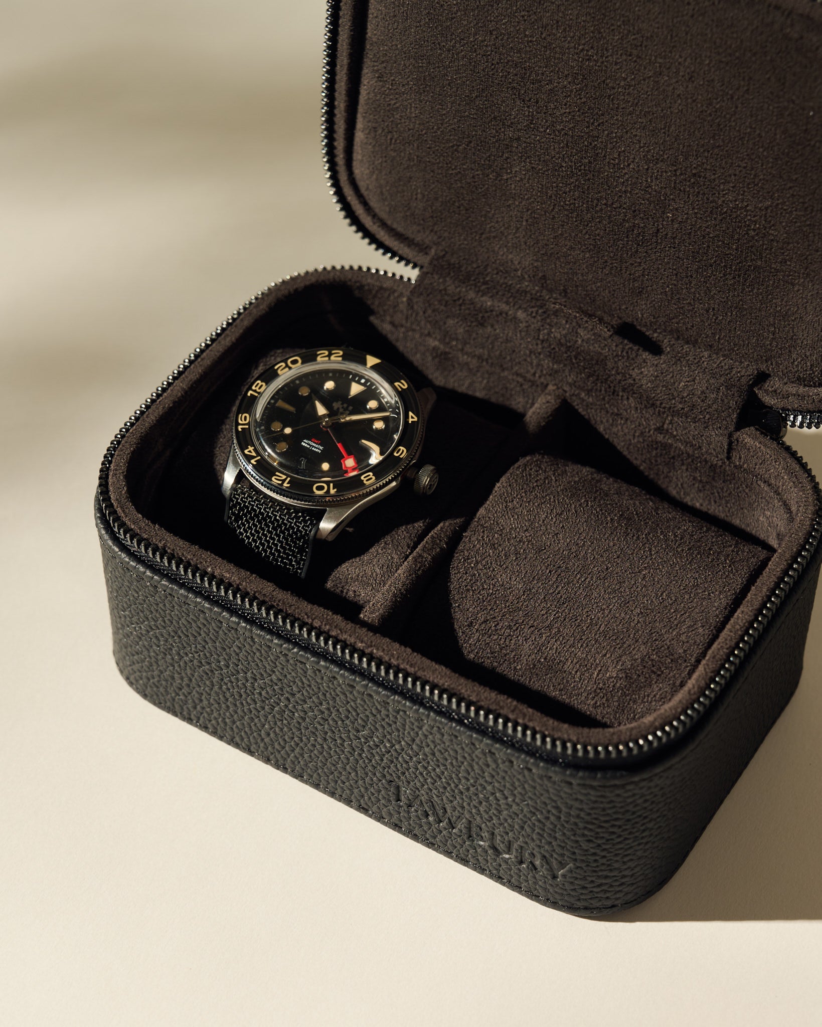 A black wristwatch with a black bezel and red accents is shown in a TAWBURY Fraser 2 Watch Case - Black, which features a textured leather design and zipper closure.
