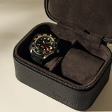 A black wristwatch with a black bezel and red accents is shown in a TAWBURY Fraser 2 Watch Case - Black, which features a textured leather design and zipper closure.