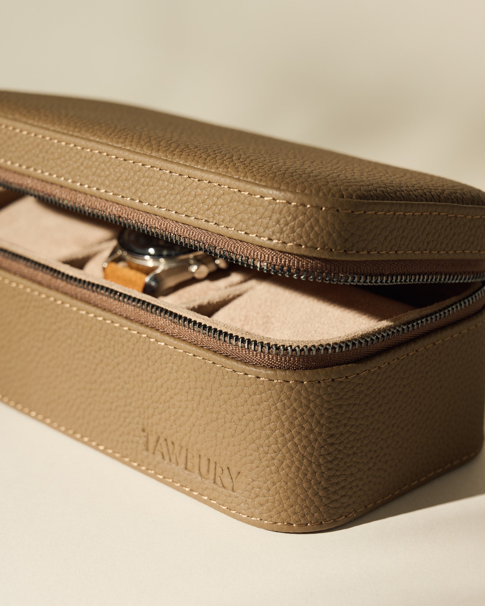 The Fraser 3 Watch Case - Taupe by TAWBURY features textured leather and a partially open zipper displaying a wristwatch inside, making it an ideal stylish storage option for your watches on the go.
