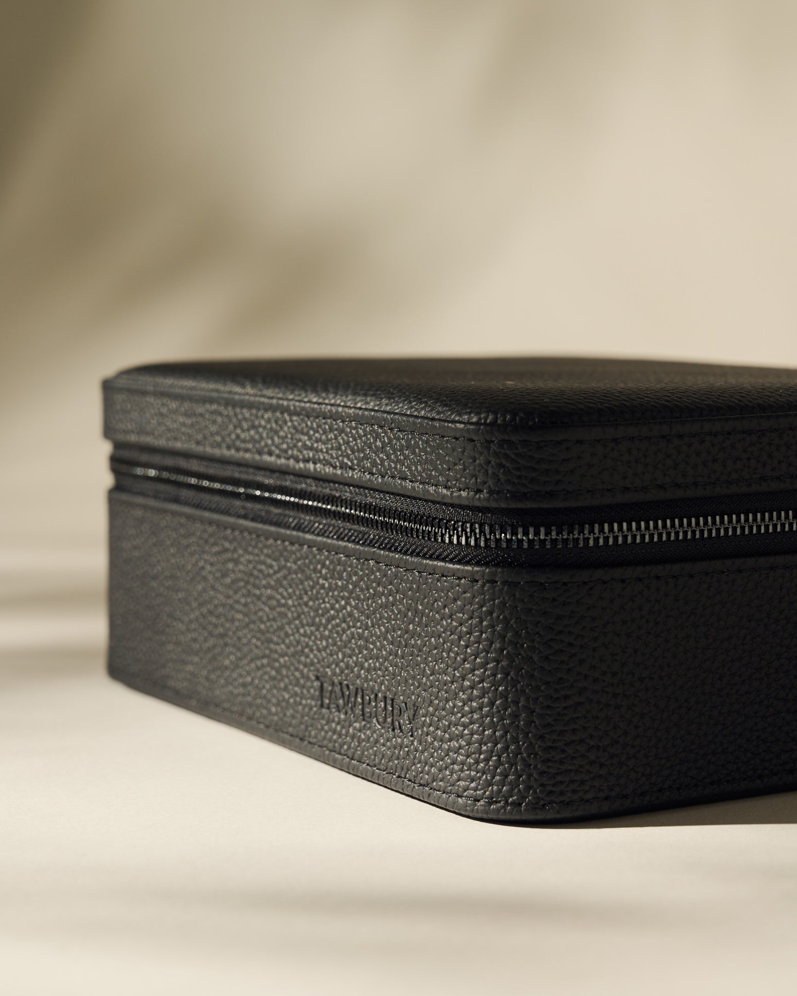 A black textured rectangular leather Fraser 3 Watch Case with Storage by TAWBURY rests on a neutral surface, featuring "TAWBURY" embossed on the side.