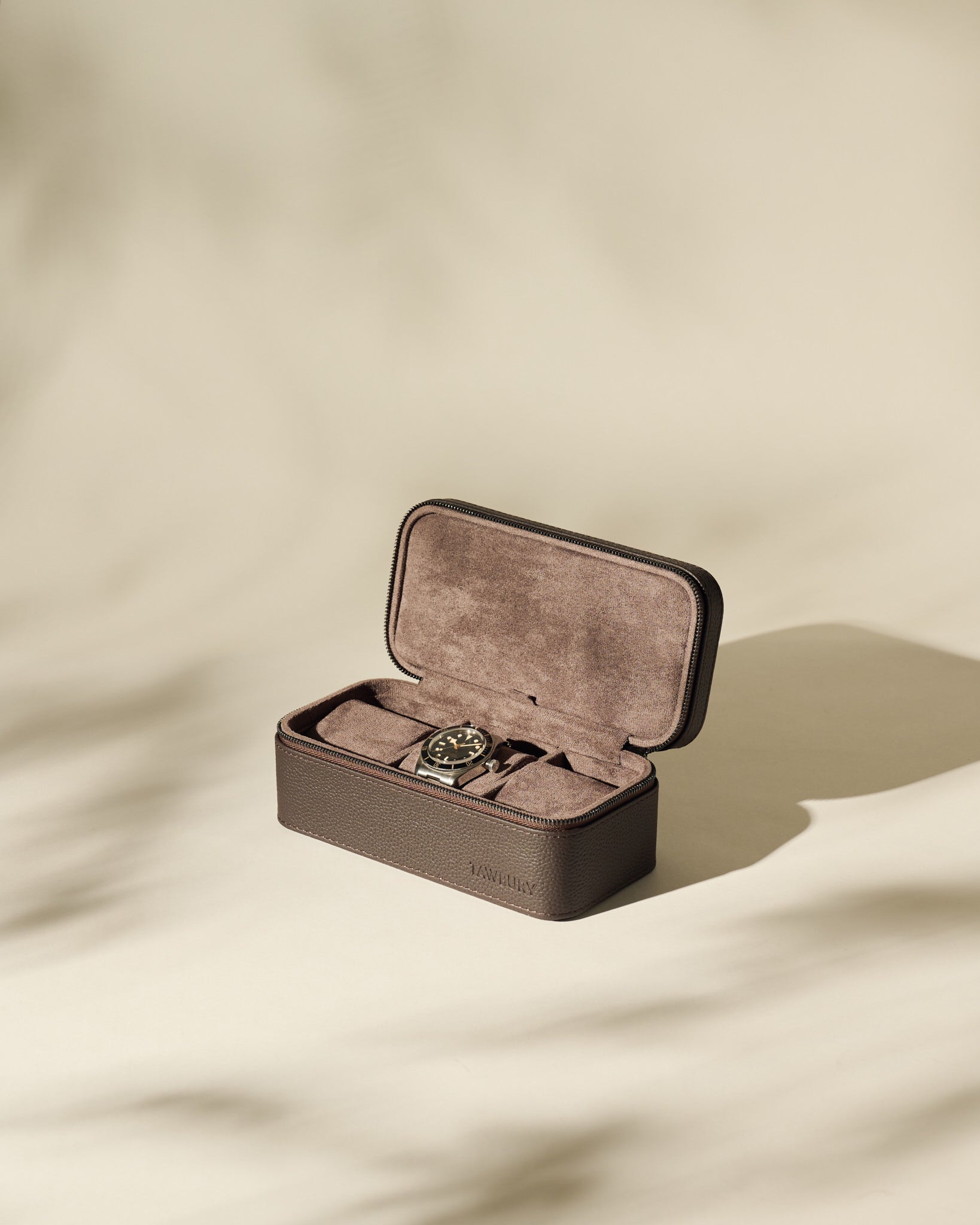 The TAWBURY Fraser 3 Watch Case in brown leather, lined with brown suede, sits open on a beige surface with soft shadows.