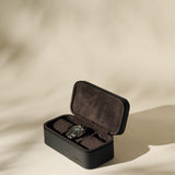 A black wristwatch rests inside the TAWBURY Fraser 3 Watch Case - Black, a sleek black and brown leather travel case, displayed on a light beige surface.