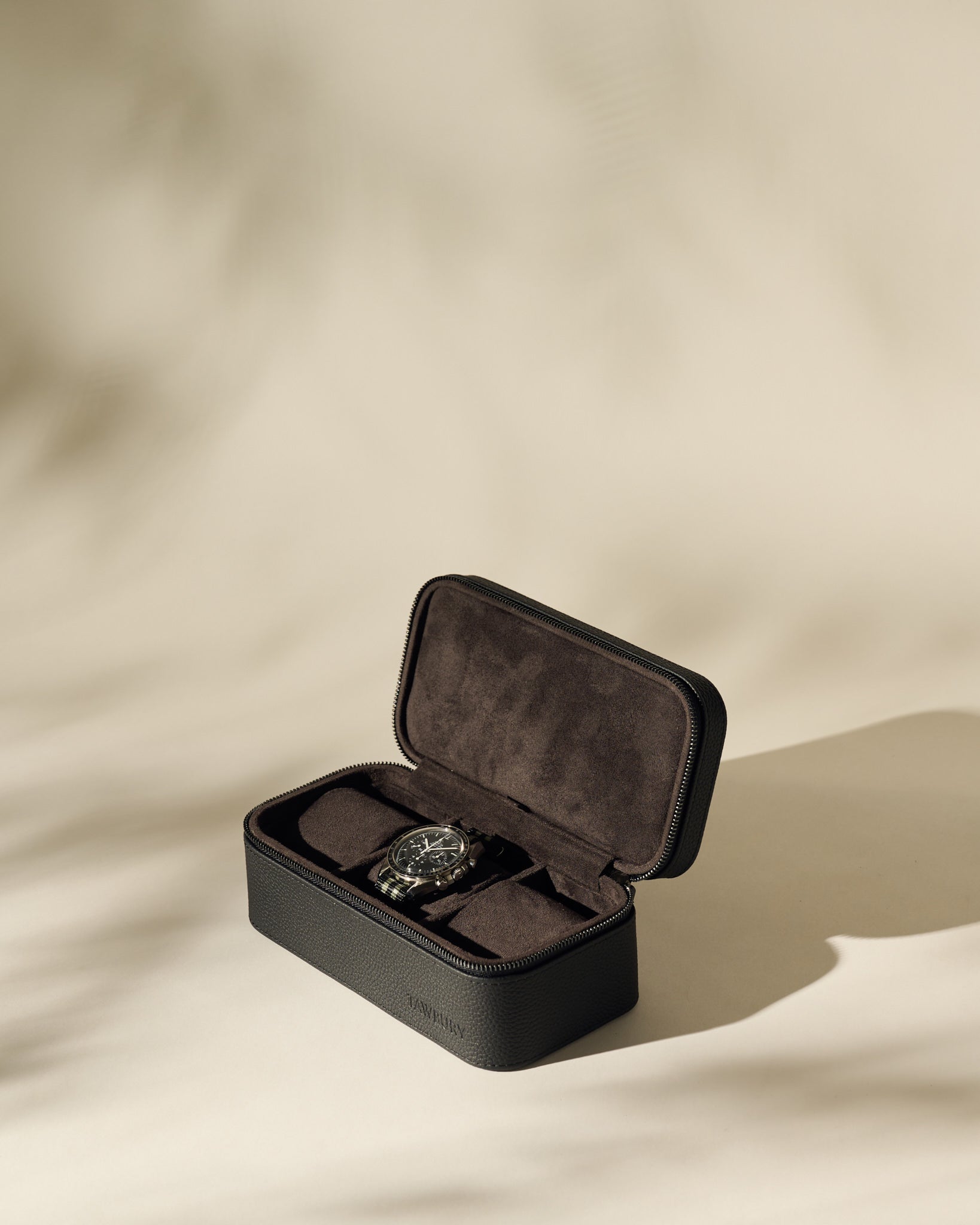 A black wristwatch rests inside the TAWBURY Fraser 3 Watch Case - Black, a sleek black and brown leather travel case, displayed on a light beige surface.