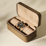 The TAWBURY Fraser 3 Watch Case in taupe features a suede interior for elegant travel storage, shown here holding a wristwatch with a black dial and brown strap in bright, neutral lighting.