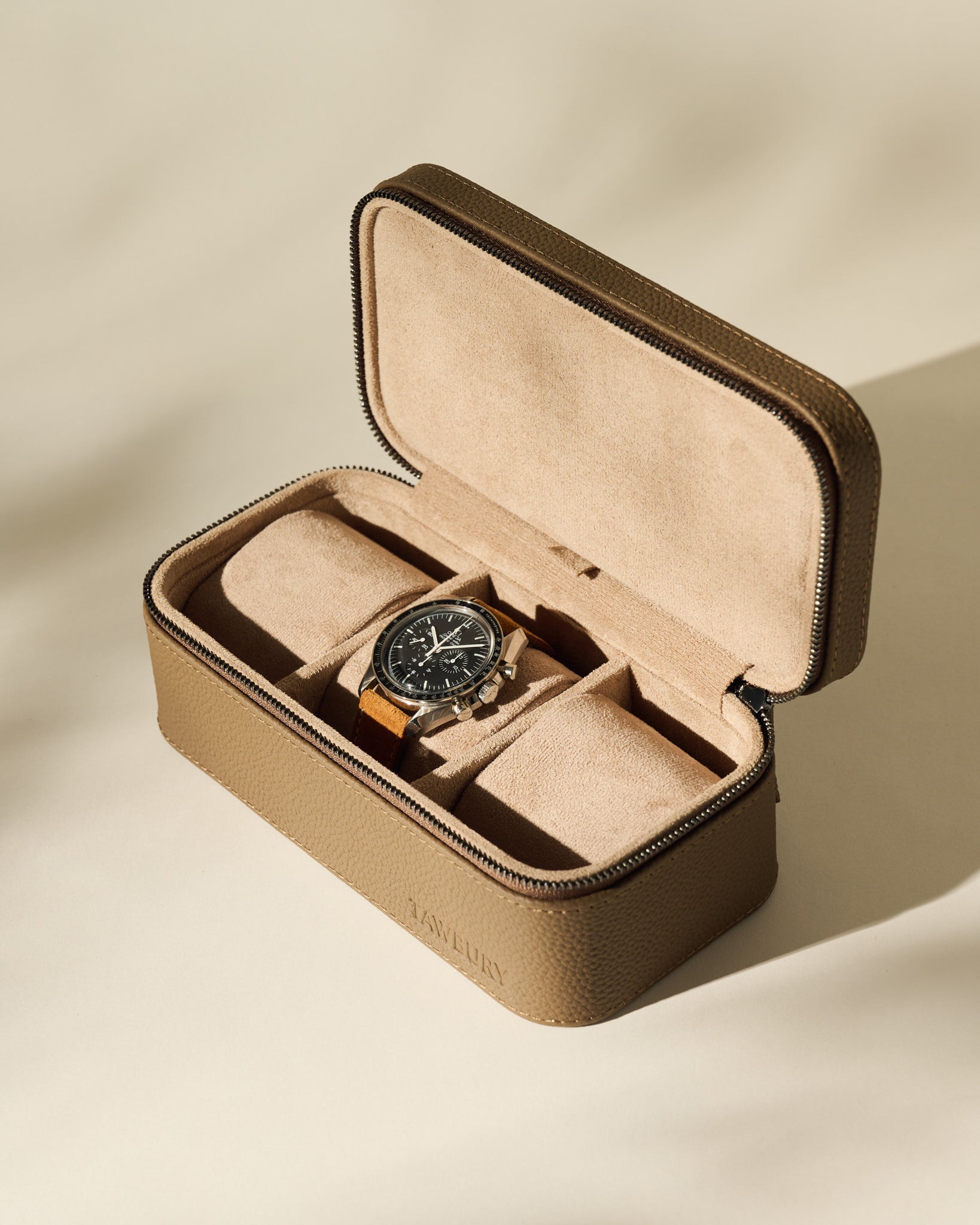 The TAWBURY Fraser 3 Watch Case in taupe features a suede interior for elegant travel storage, shown here holding a wristwatch with a black dial and brown strap in bright, neutral lighting.