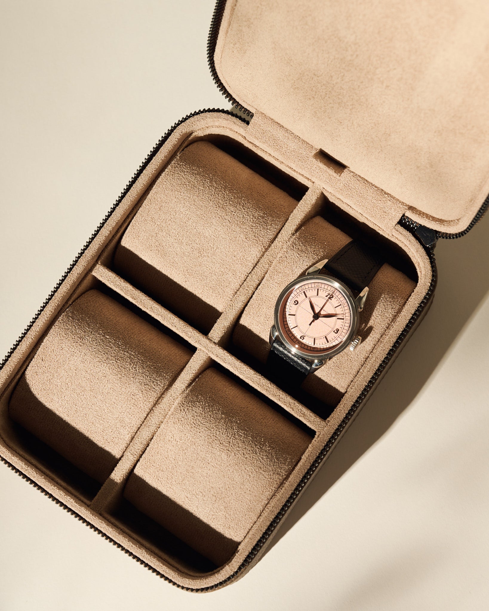 A black leather watch with a pink face sits in the TAWBURY Fraser 4 Watch Case - Taupe, which features tan suede lining and four compartments, with one occupied.