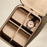 A black leather watch with a pink face sits in the TAWBURY Fraser 4 Watch Case - Taupe, which features tan suede lining and four compartments, with one occupied.
