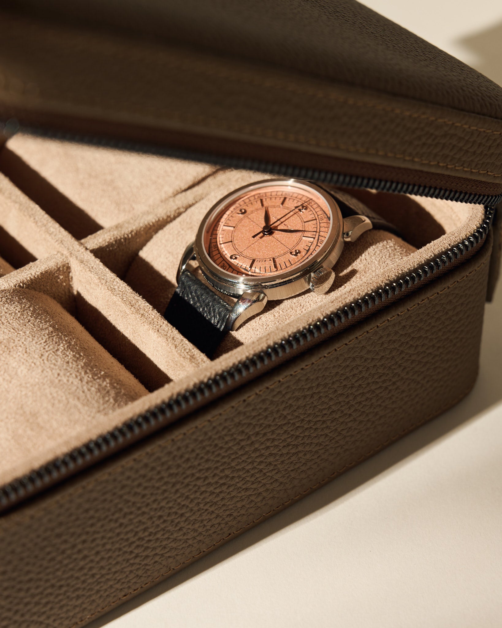 A TAWBURY Fraser 4 Watch Case in taupe, featuring a soft, suede-like interior and secure zipper closure, stores a wristwatch with a black leather strap and rose gold face.