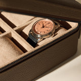 A TAWBURY Fraser 4 Watch Case in taupe, featuring a soft, suede-like interior and secure zipper closure, stores a wristwatch with a black leather strap and rose gold face.