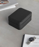 A black leather zippered TAWBURY Fraser 4 Watch Travel Case - Black (Coming Soon) is placed on a gray surface, next to an open magazine and a wristwatch with a stainless steel band.