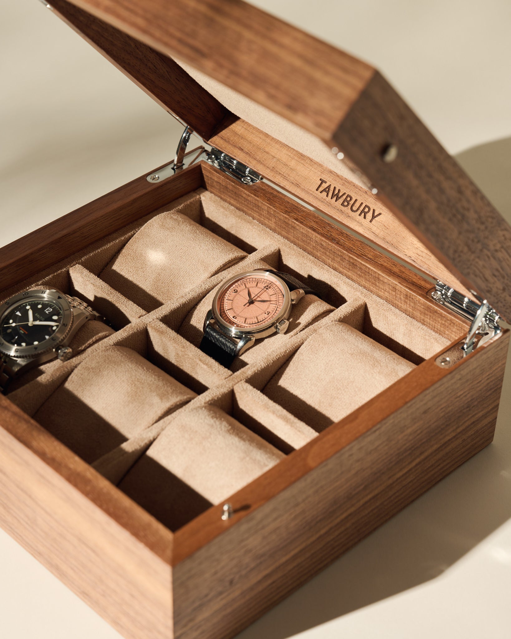 A TAWBURY Grove 6 Slot Wooden Watch Box in light walnut with a solid lid shows two wristwatches on beige cushioned slots, while the other compartments are empty.