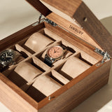 A TAWBURY Grove 6 Slot Wooden Watch Box in light walnut with a solid lid shows two wristwatches on beige cushioned slots, while the other compartments are empty.