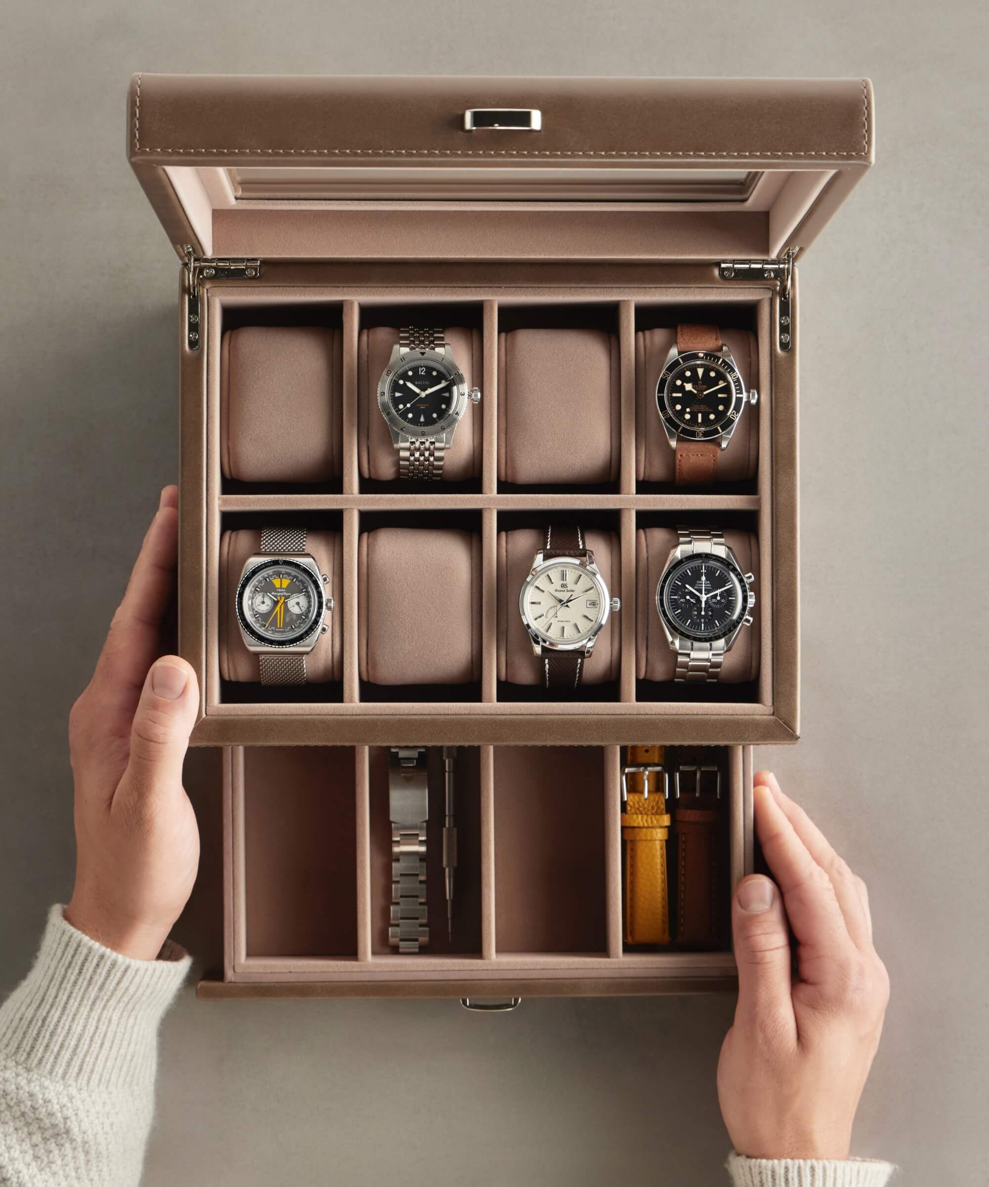 A watch enthusiast's dream: hands revealing a TAWBURY Bayswater 8 Slot Watch Box with Storage in taupe, elegantly showcasing six watches alongside additional straps crafted from vegan leather.