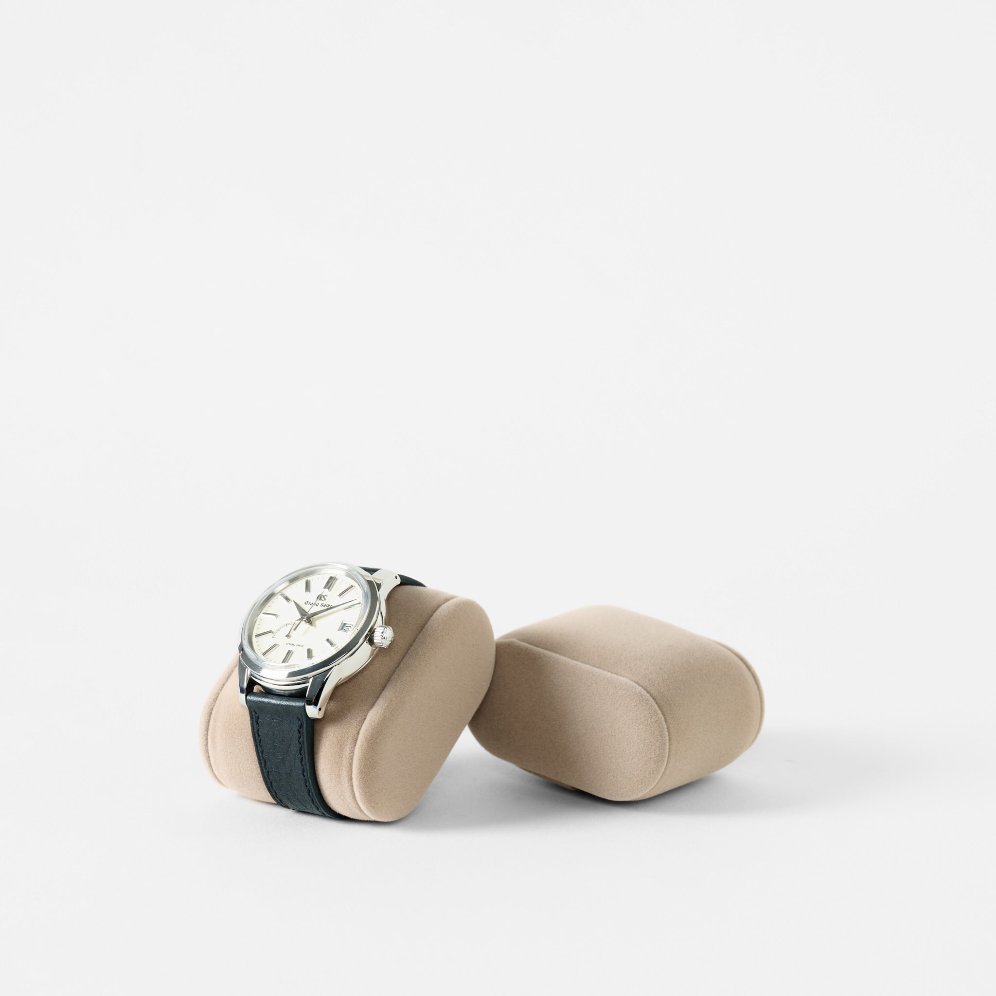 A wristwatch with a black strap rests on a TAWBURY Bayswater Watch Pillow - Beige - 55mm - Small, next to an identical empty cushion, both displayed against a plain white background.