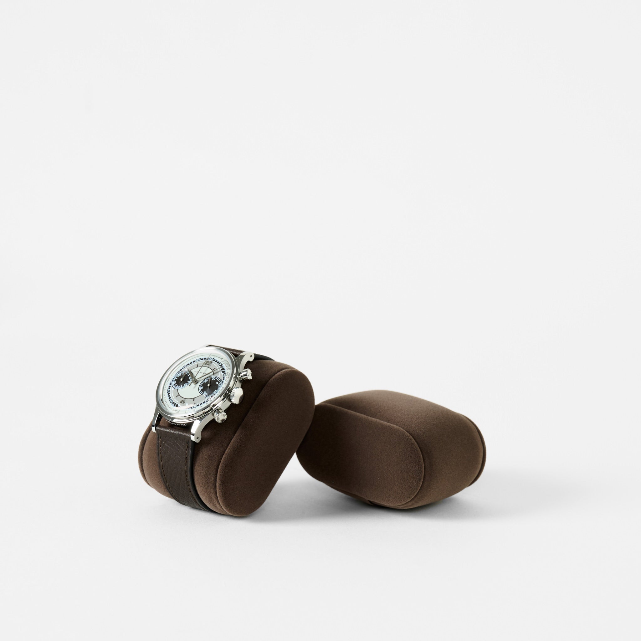 A silver watch with a black strap sits on a brown TAWBURY Bayswater Watch Pillow (50mm, Small), beside an empty matching cushion—both Brown/Grey pillows are shown against a plain white background.