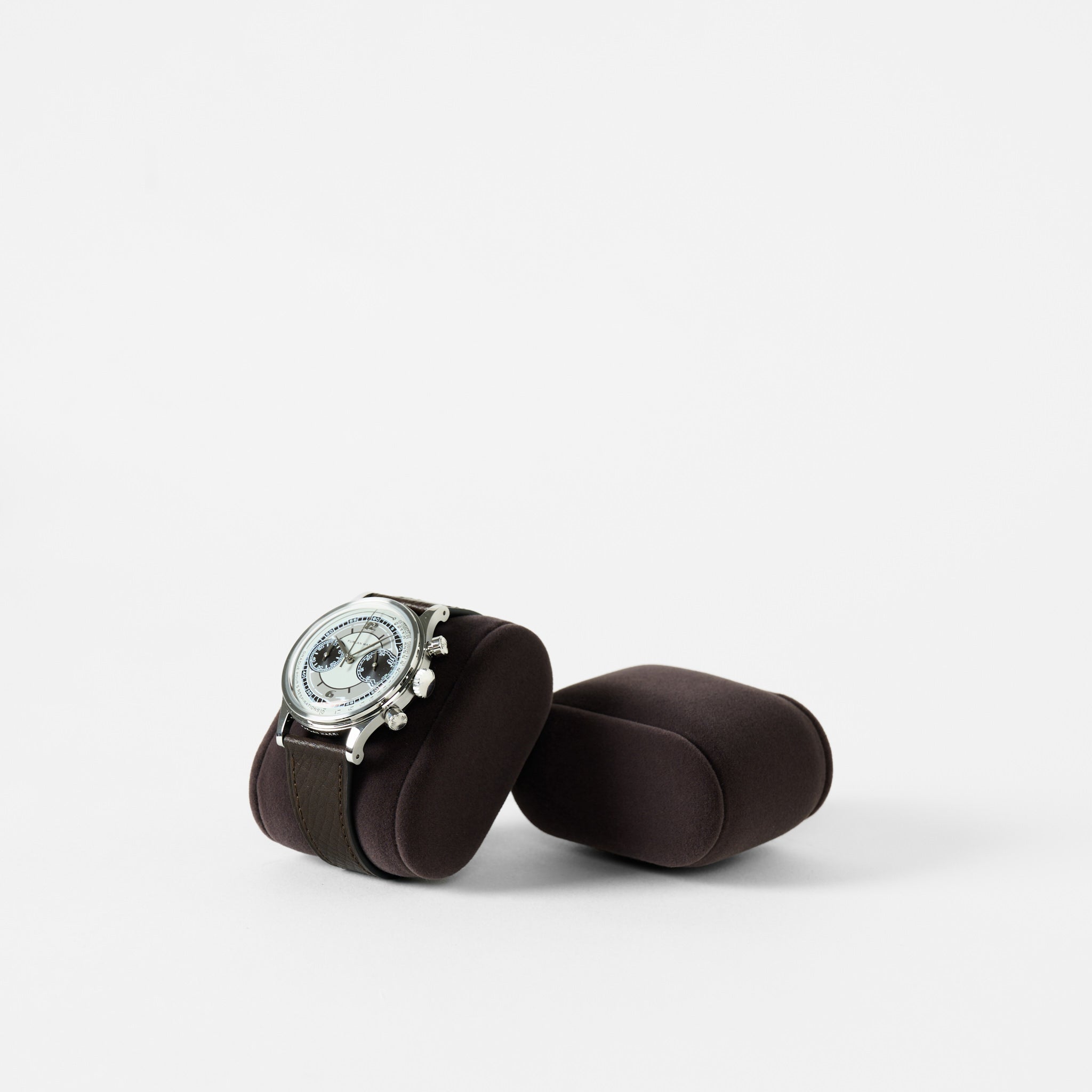 A TAWBURY Bayswater Watch Pillow in Charcoal (50mm, Small) displays a silver wristwatch with a dark leather strap on a brown cushioned stand, with an identical empty stand nearby against a plain white background.
