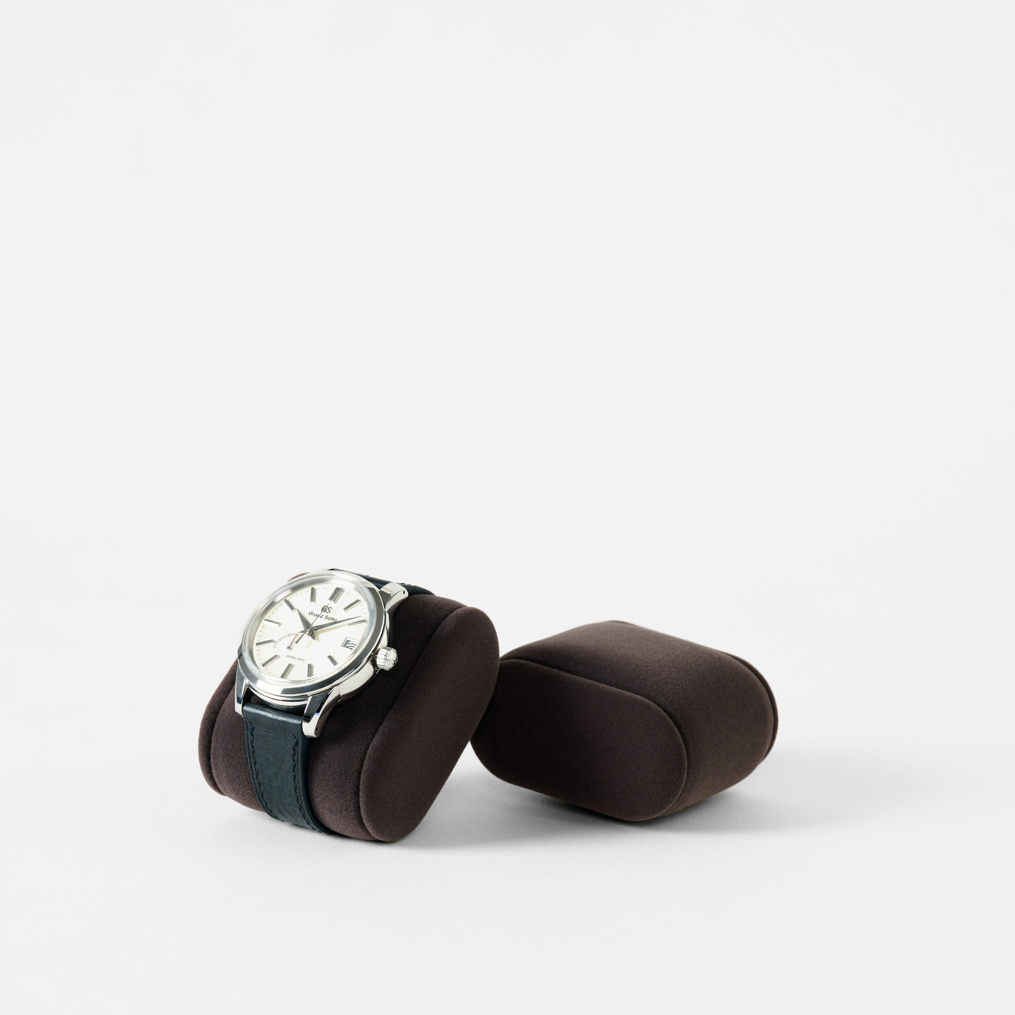 A silver wristwatch with a black strap rests on a TAWBURY Bayswater Watch Pillow in Charcoal (55mm, Small), beside an empty matching pillow, both displayed against a plain white background.
