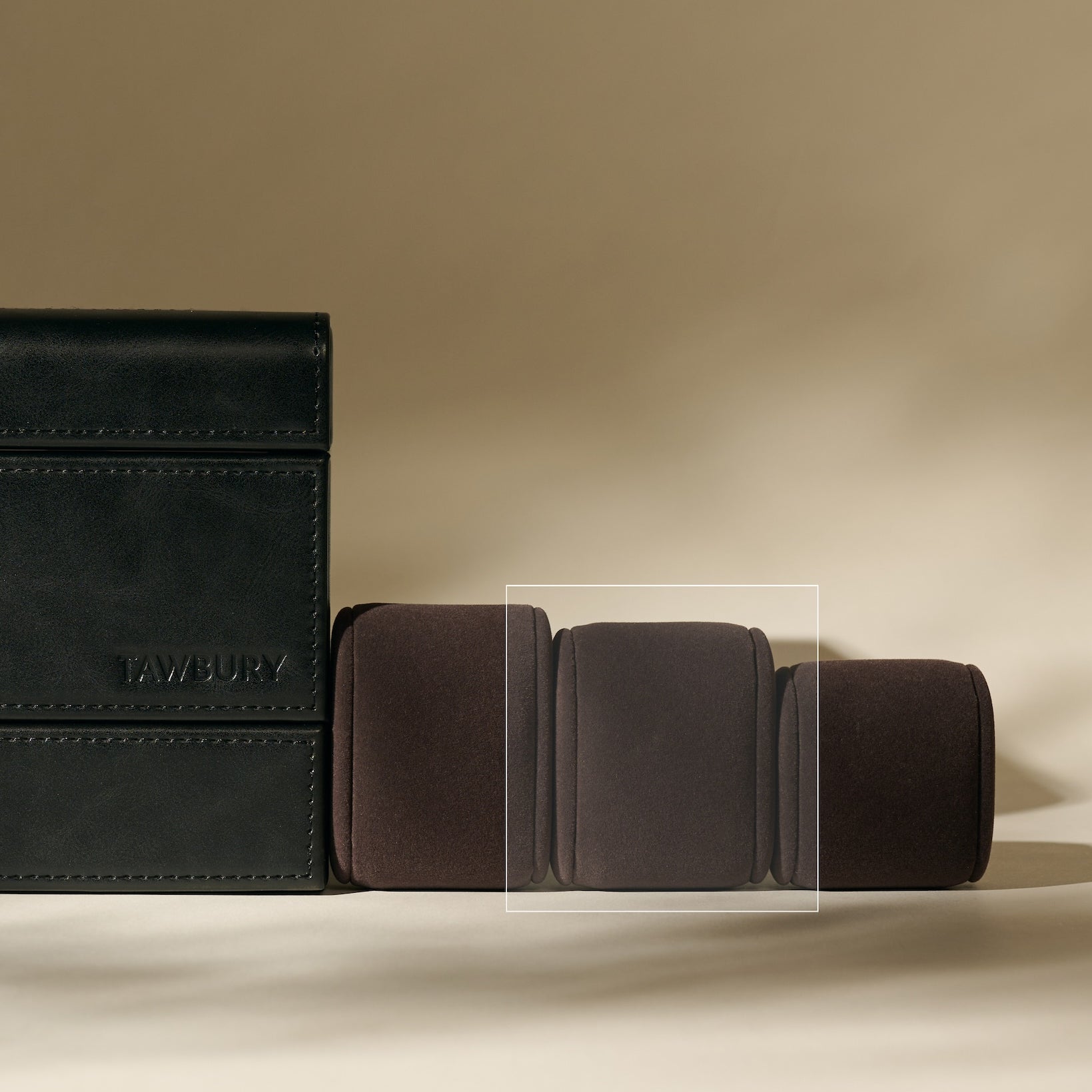 A black leather wallet sits by three closed brown leather wallets on a beige backdrop, alongside TAWBURY Bayswater Watch Pillows in Charcoal (55mm, Small)—ideal for slender wristwatches.