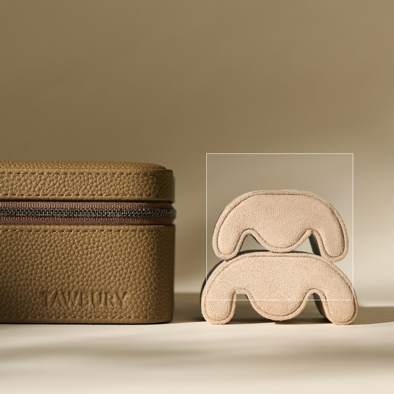 A textured brown pouch with a zipper sits beside two stacked TAWBURY Fraser Watch Pillows (X-Small, Beige), shaped abstractly and suede-like, against a neutral background.