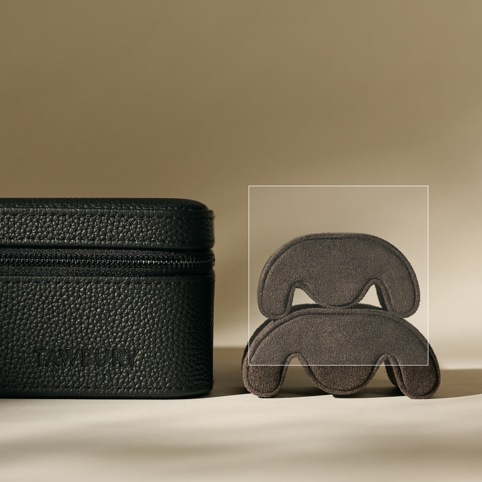 A set of TAWBURY Fraser Watch Pillows - X-Small in Anthracite are arranged on a beige surface beside a sculptural gray object, their sleek design complementing the neutral background.