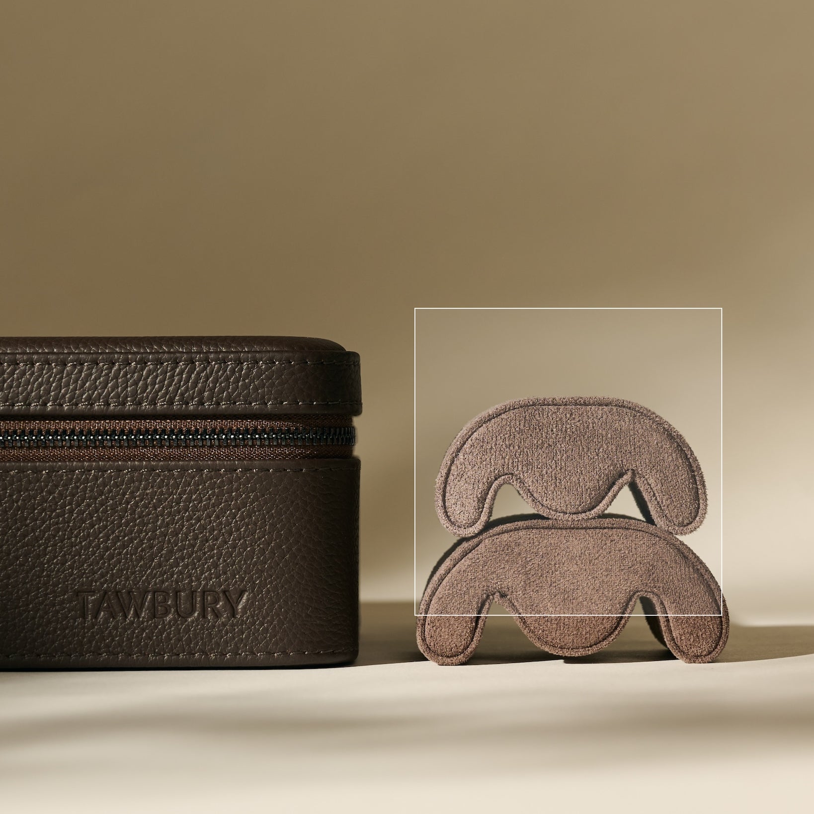 A TAWBURY Fraser Watch Travel Case with a zipper sits beside two stacked TAWBURY Fraser Watch Pillows - X-Small - Brown, all set on a beige background.