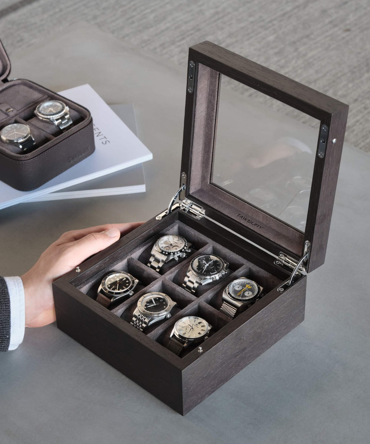 A person's hand is near an open TAWBURY Grove 6 Slot Wooden Watch Box with Kassod wood veneer and a glass lid, containing six watches arranged in compartments. The box is placed on a table next to a small closed case with additional watches and a closed book.