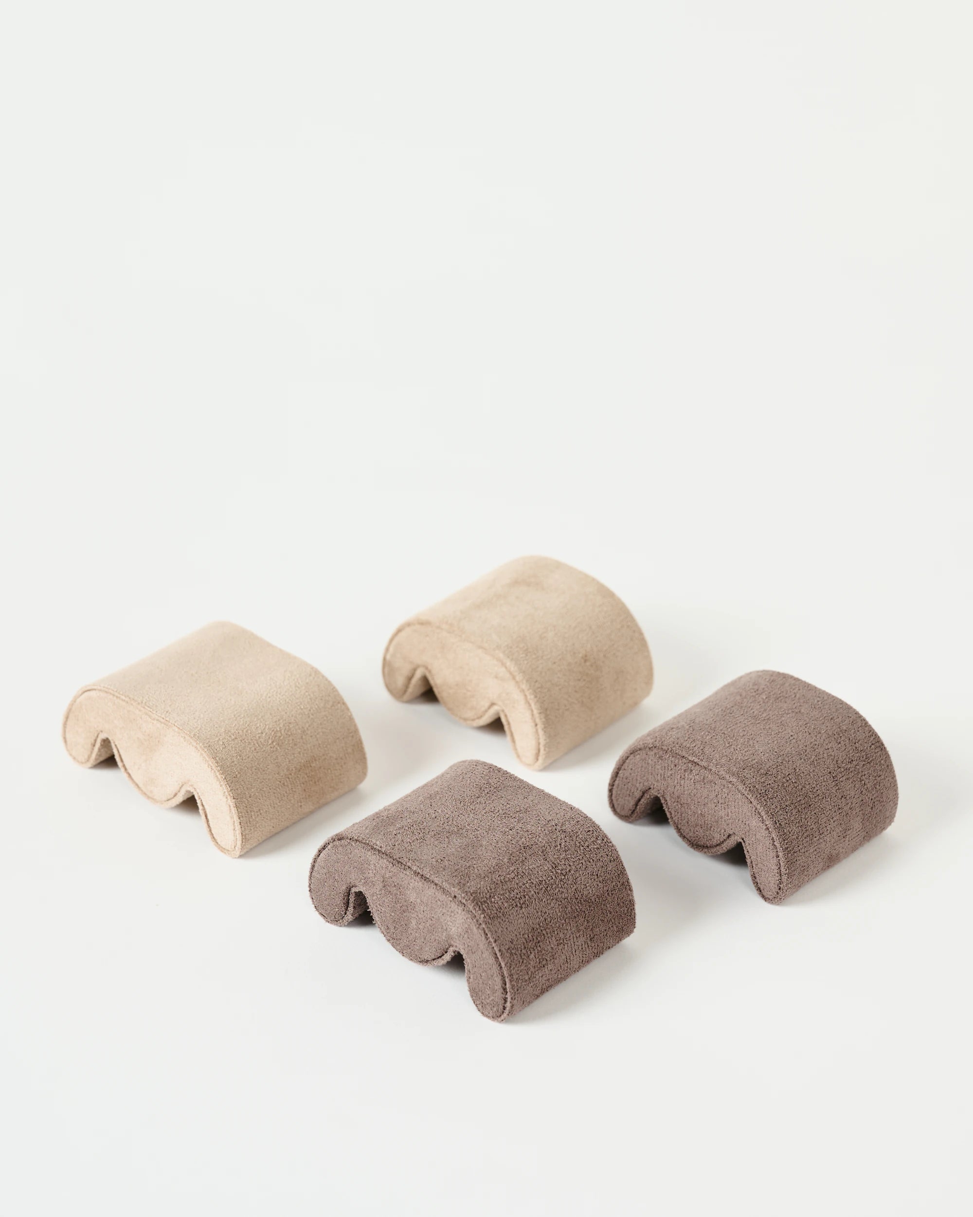 Four soft, curved cushion pads in light beige and brown sit on a white surface, ideal for organizing your timepieces in the TAWBURY Grove 6 Slot Wooden Watch Box—Dark Walnut Wood with Glass Lid.