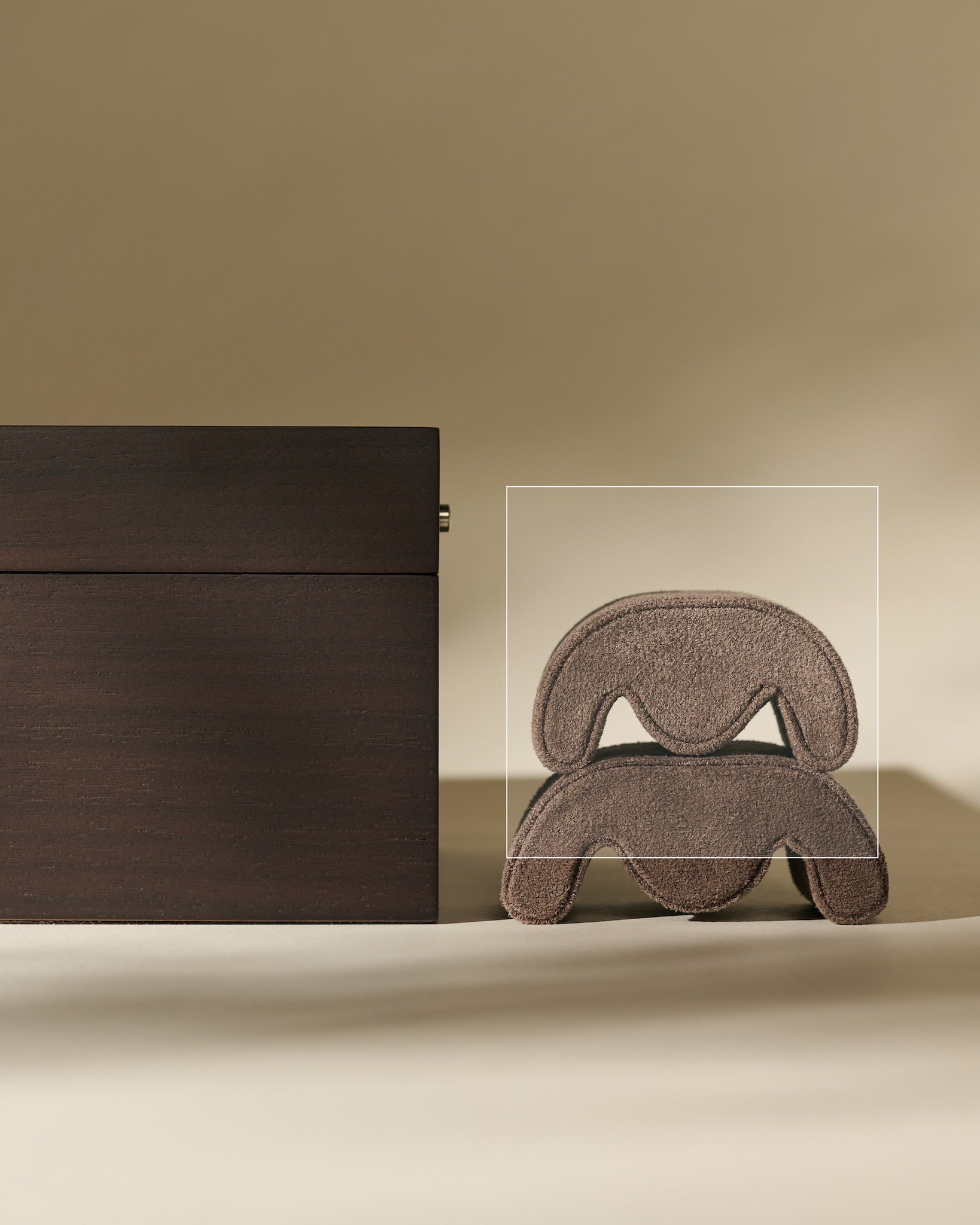 A set of TAWBURY Grove Watch Pillows - X-Small - Brown rests in a rectangular box beside a sculptural, layered brown piece, all on a beige background—ideal for supporting your watch or fitting a slender wrist.