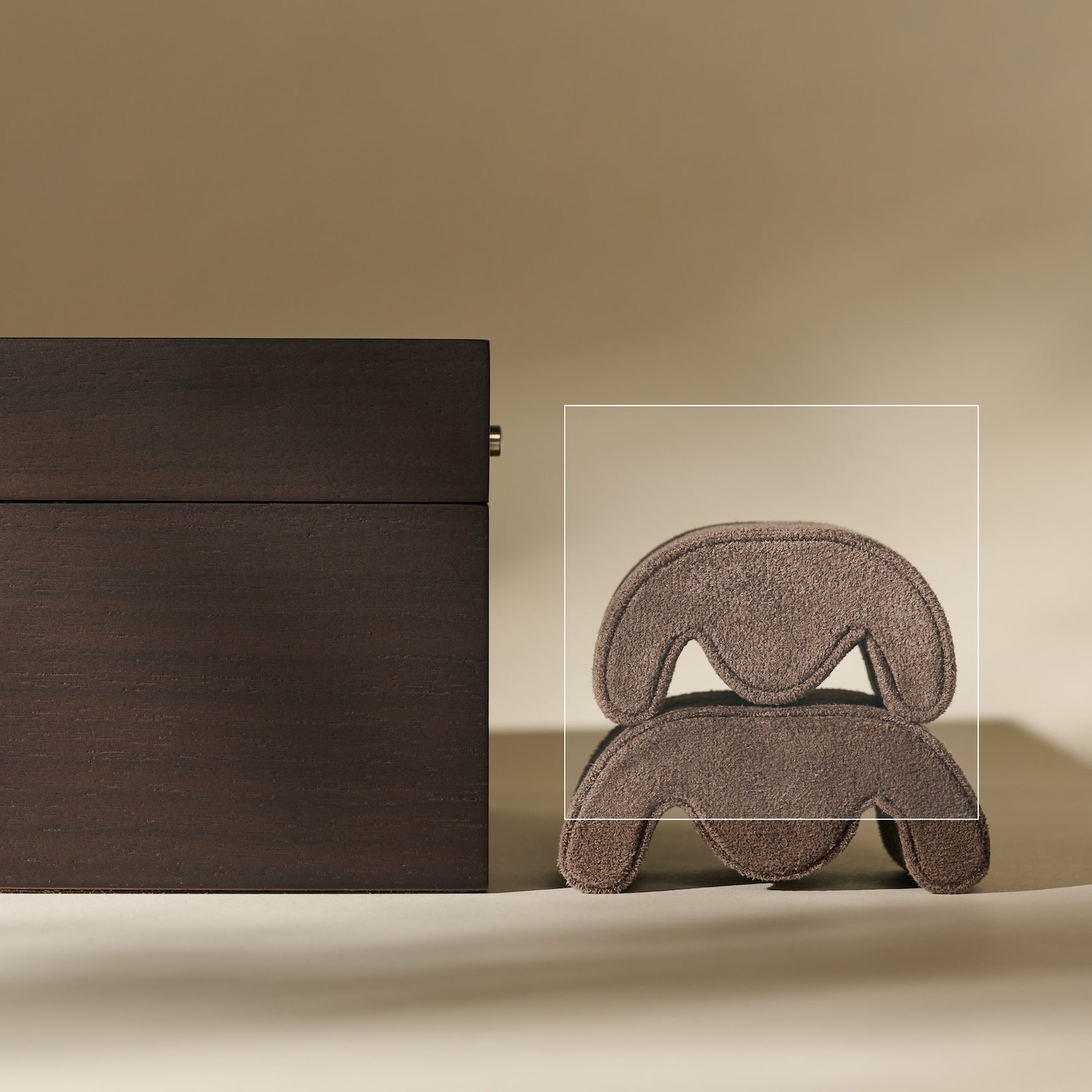 A set of TAWBURY Grove Watch Pillows - X-Small - Brown rests in a rectangular box beside a sculptural, layered brown piece, all on a beige background—ideal for supporting your watch or fitting a slender wrist.