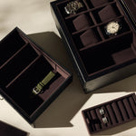 Three Bayswater 6 Watch Jewelry Boxes by TAWBURY in black/charcoal are displayed on a beige surface: one holds two wristwatches, another is a vegan leather box with an olive green strap, and the third contains metal watch links.