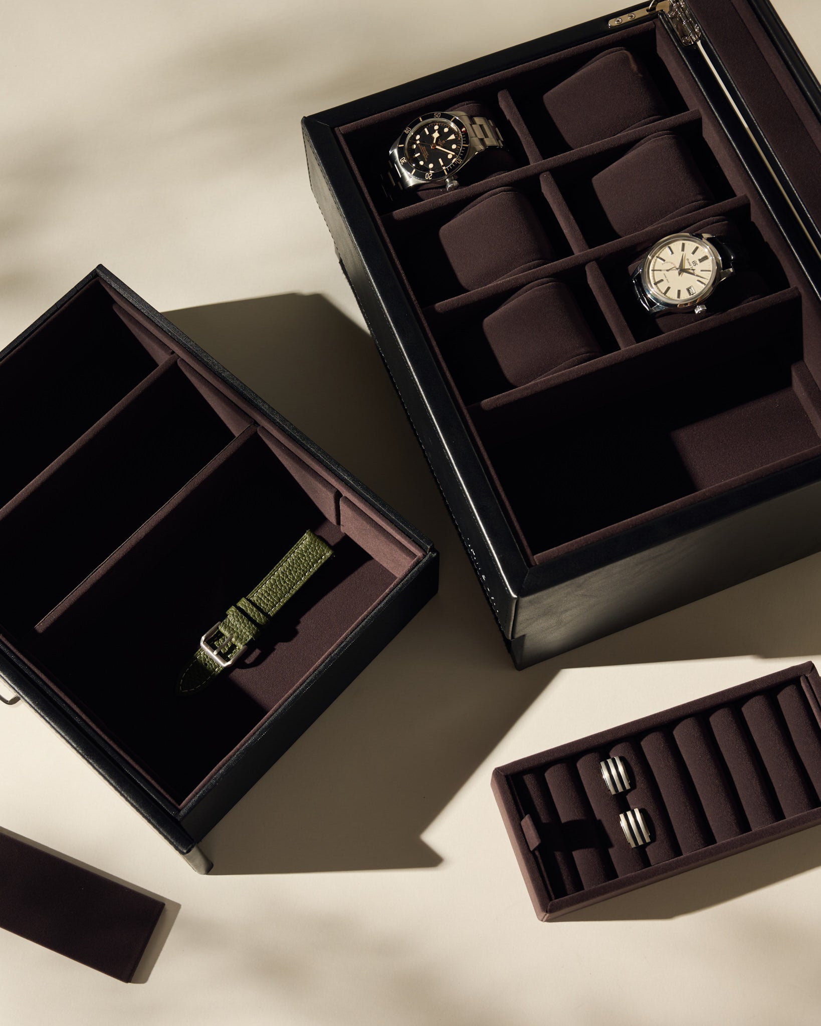 Three Bayswater 6 Watch Jewelry Boxes by TAWBURY in black/charcoal are displayed on a beige surface: one holds two wristwatches, another is a vegan leather box with an olive green strap, and the third contains metal watch links.