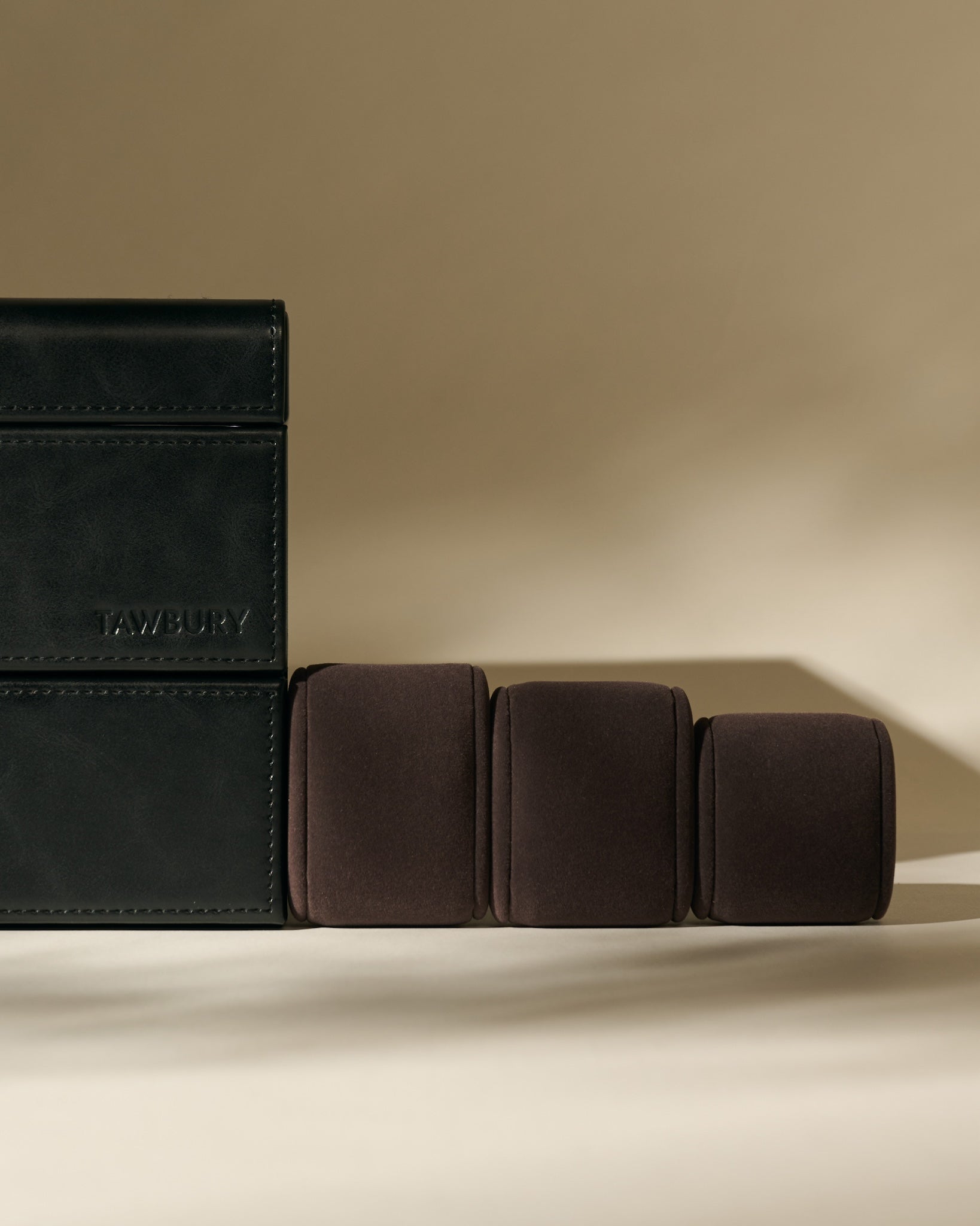 The TAWBURY Bayswater 6 Watch Jewelry Box in midnight black is displayed beside three brown velvet watch pillows on a beige background.