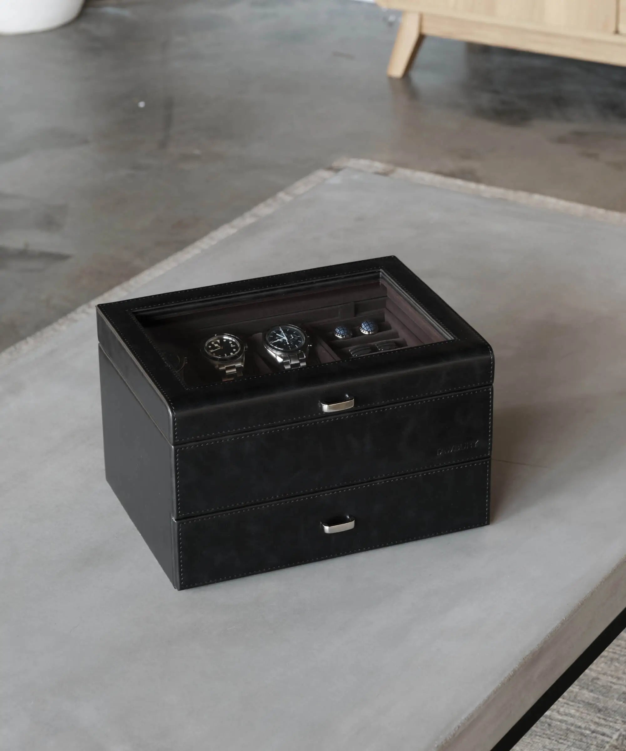 A TAWBURY Bayswater 6 Watch Jewellery Box in Black/Charcoal with a glass top showcases six watches and extra storage, elegantly placed on a light gray table in a modern room.