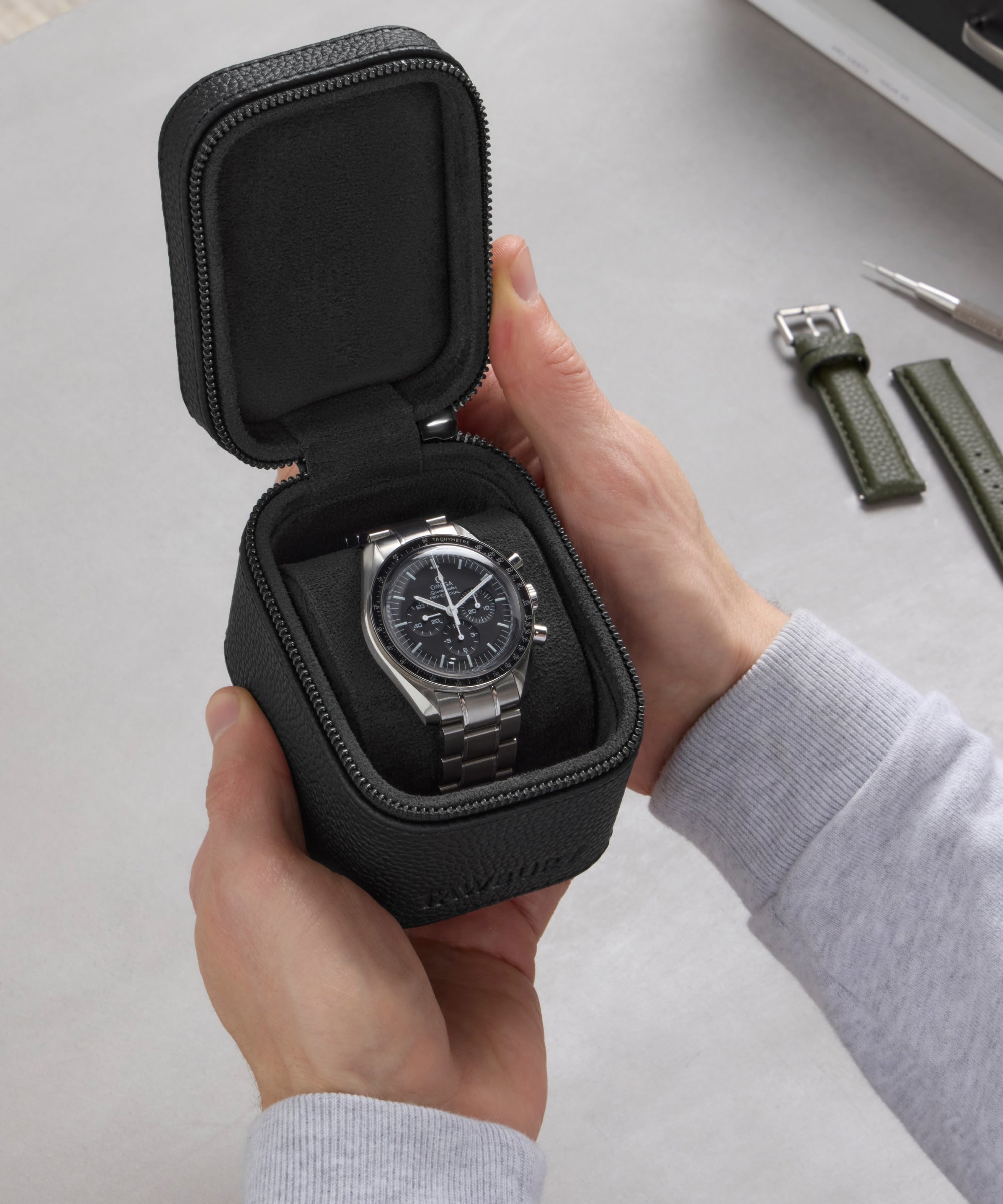 A person holds the TAWBURY Fraser 1 Watch Travel Case - Black (Coming Soon), featuring a stainless steel wristwatch with a green strap inside, while a tool rests on the table—ideal for safe watch travel.