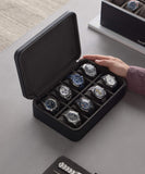 A person opens the Fraser 8 Watch Travel Case - Black from TAWBURY, revealing assorted wristwatches organized in separate compartments on a sleek gray surface.