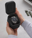 A person holds the TAWBURY Fraser 1 Watch Travel Case - Black (Coming Soon), featuring a stainless steel wristwatch with a green strap inside, while a tool rests on the table—ideal for safe watch travel.