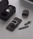 The Fraser 3 Watch Travel Case by TAWBURY elegantly showcases two watches on black pillows, one protected in a leather case, alongside a green strap, extra clasp, and box lid—all arranged on a gray surface—perfect for your next adventure.
