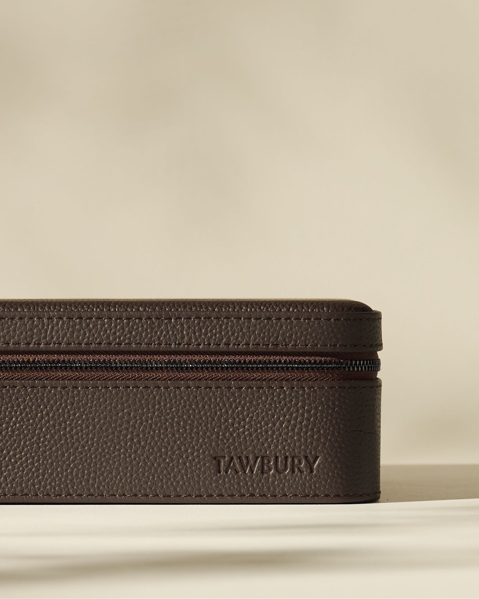 The Fraser 6 Watch Case by TAWBURY features brown textured leather, a zippered closure, and “TAWBURY” embossed on the side, displayed against a neutral background.