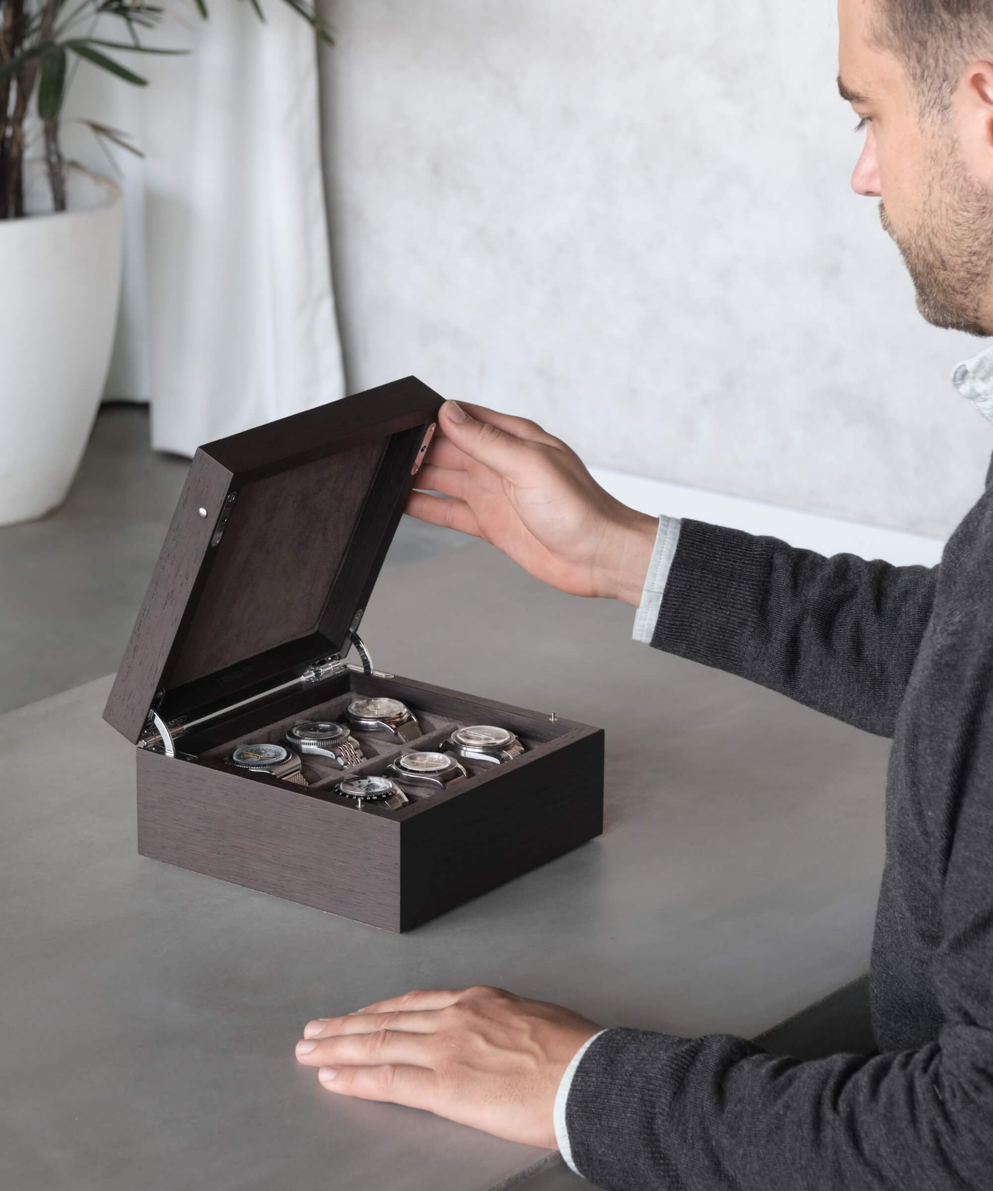 A man opening a TAWBURY Grove 6 Slot Watch Box with Solid Lid - Kassod on a table, revealing a collection of elevate timepieces.