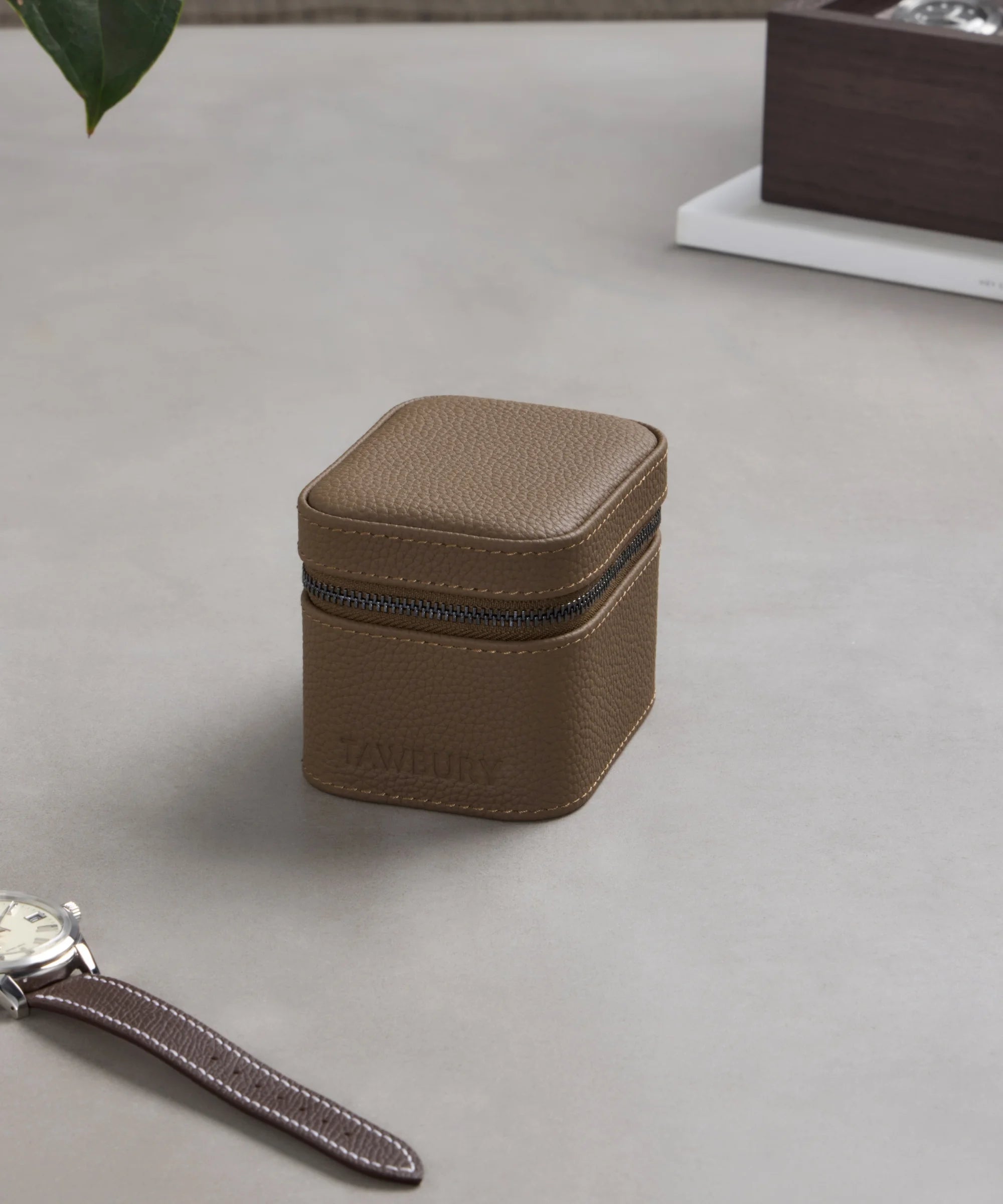 The TAWBURY Fraser 1 Watch Travel Case in taupe sits on a light gray surface beside a wristwatch with a matching brown leather strap.