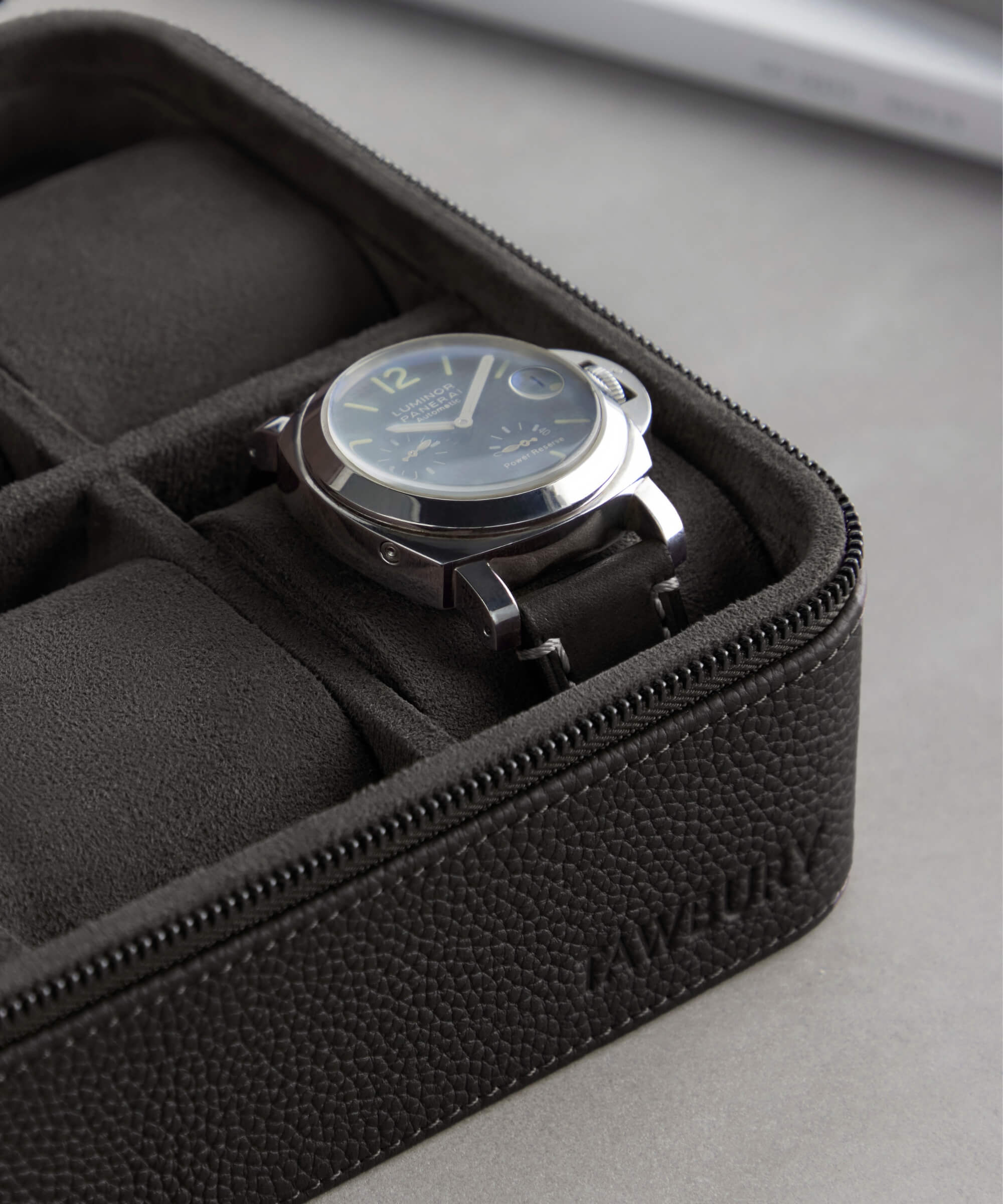 A wristwatch with a black band rests in a sleek leather watch case with a partially unzipped lid and the brand name "TAWBURY" embossed on the case, part of the distinguished Fraser 4 Watch Travel Case - Black (Coming Soon) collection.