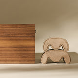 The TAWBURY Grove 6 Slot Wooden Watch Box in classic walnut wood with a glass lid stands beside two stacked, curved beige objects resembling abstract stools or cushions, set against a neutral background.