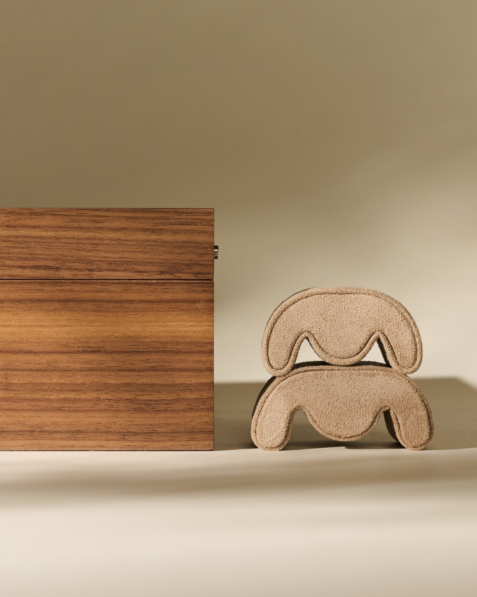 The TAWBURY Grove 6 Slot Wooden Watch Box in classic walnut wood with a glass lid stands beside two stacked, curved beige objects resembling abstract stools or cushions, set against a neutral background.
