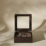A TAWBURY Grove 6 Slot Wooden Watch Box in Dark Walnut with a glass lid is open, stylishly presenting two wristwatches inside against a beige background.